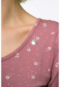 Rosa Top mit Rundhalsausschnitt und weißem Blumenmuster, mit einem silbernen, runden Akzent am Kragen. Hergestellt aus weichem, strukturiertem Stoff.