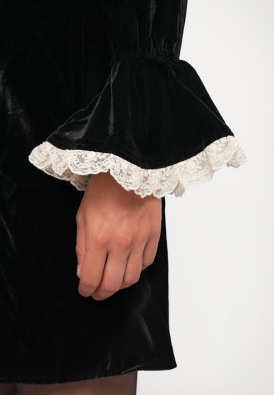 Black velvet garment with flared sleeves. Lace trim on cuffs in off-white. Hand visible holding the fabric. Smooth texture, elegant design.