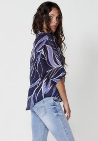 Camicia a maniche corte blu e viola a motivi, con una vestibilità rilassata, abbinata a jeans blu chiaro. Il tessuto sembra leggero e tridimensionale.