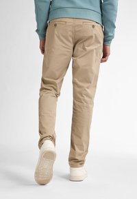 Beige katoenen broek met een slim fit, twee achterzakken met knopen en een taps toelopend design. G gedragen met witte sneakers.