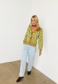 Ξανθιά γυναίκα που φοράει πράσινη ζακέτα με βούλες και πορτοκαλί λεπτομέρειες, ανοιχτό μπλε τζιν, καφέ τακούνια με καρφιά και φουλάρι με σχέδια στο λαιμό, στέκεται σε εσωτερικό χώρο.