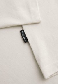 Tissu blanc avec une étiquette tissée noire portant l'inscription "LEVEL UP ombre" cousue dans une couture entre deux couches superposées.