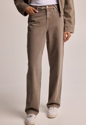 Beige Hosen mit geradem Schnitt und dünnem Gürtel, kombiniert mit weißen Turnschuhen und einer teilweise sichtbaren beigen Jacke, getragen von einer stehenden Person.