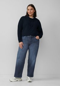 Frau steht da, trägt einen navy-blauen Hoodie, blue straight-leg Jeans und weiße Sneaker, mit einer Hand in der Tasche vor einem schlichten grauen Hintergrund.