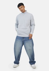 Felpa grigia con zip e colletto alto, abbinata a jeans in denim blu larghi e sneakers bianche.