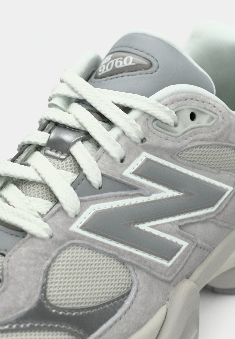 Grauer und weißer Sportschuh mit dicken weißen Schnürsenkeln, Mesh- und Wildlederelementen sowie einem "9060"-Etikett auf der gepolsterten Zunge.