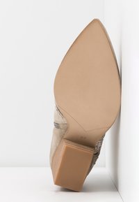 Beige semišové ankle boty s špičatou špičkou a blokovým podpatkem, s podrážkou směřující dopředu, velikost 37 viditelná, umístěné proti bílým rohovým stěnám.