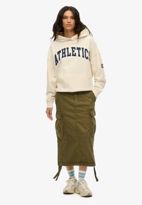 Crème hoodie met marineblauwe tekst "ATHLETICS", kengaroo zak; olijfgroene cargorok met zakken, verstelbare zijstrikken; witte sneakers met blauwe accenten.