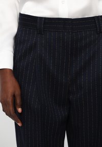 Navy pinstripe trousers made from a textured fabric, featuring a flat front, traditional waistband, and side pockets.