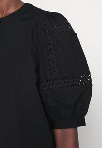 Manche de blouse noire avec un design bouffant et des détails en dentelle florale découpée sur le haut du bras et le poignet, portée par une personne à la peau foncée.