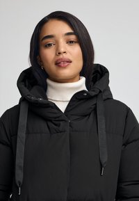 Svart pufferjacka med huva, med dragkedjestängning, texturerad tyg och logotypprägling på snörena. Bärs över en vit polotröja.