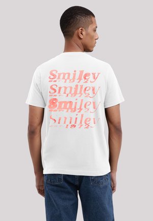 Weißes Baumwoll-T-Shirt mit rotem verzerrtem "Smiley"-Text und der Jahreszahl "1972" auf der Rückseite. Kurze Ärmel und eine lockere Passform.