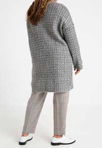 Lång, oversized grå tröja med ett strukturerat stickat mönster, tillsammans med rutiga byxor och vita skor. Löst sittande med ärmar som når till armbågarna.