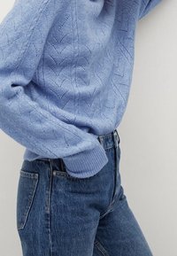 Světle modrý vzorovaný svetr zastrčený do tmavě modrých džín s vysokým pasem, s jednou rukou částečně v přední kapse.