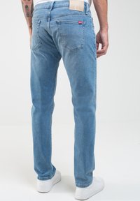 Vaqueros de denim azul claro con un corte recto. Cuenta con cinco bolsillos y un parche de cuero en la parte trasera de la cintura. Se lleva con zapatillas blancas.