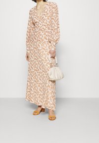 Blumiges Maxikleid in Creme mit braunem Blattmuster, ausgestattet mit langen Ärmeln und einem tiefen V-Ausschnitt. Kombiniert mit einer weißen Handtasche und gelben Sandalen.