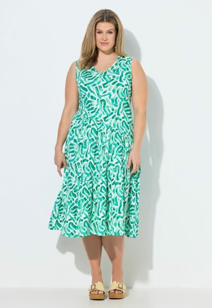 Kobieta stoi w bezrękawowej sukience midi z zielono-turkusowym abstrakcyjnym wzorem i beżowymi sandałami na platformie na białym tle.