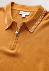 Polo en maille jaune moutarde avec double rayure blanche sur le col et fermeture éclair argentée partiellement ouverte.