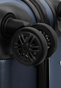 Gros plan sur des doubles roues noires pivotantes attachées à une valise rigide texturée bleue près de la fermeture éclair.