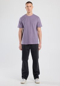 Junger Mann steht, trägt ein schlichtes lila T-Shirt mit kurzen Ärmeln, schwarze Jeans und weiße Sneaker vor einem schlichten grauen Hintergrund.