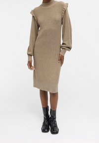 Femme portant une robe midi en tricot côtelé beige avec des détails de poignets volantés et de longues manches bouffantes, associée à des bottines noires en cuir verni.
