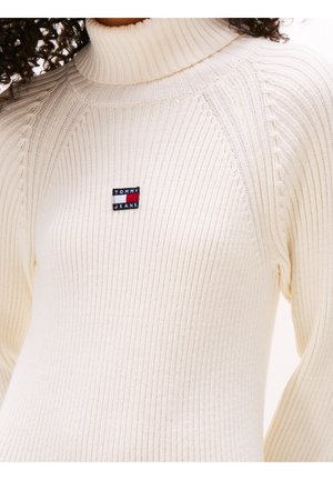 Cream-colored ribbed turtleneck sweater with small red, white, and blue Tommy Jeans logo patch on chest.