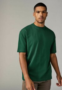 Camiseta de algodón verde de manga corta con cuello redondo, de corte holgado y textura suave. Llevada con pantalones marrón claro.