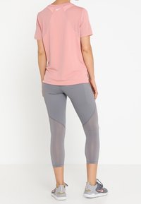 Helles rosa Sport-T-Shirt mit Netzpaneelen und grauen Leggings, die eine schlanke Passform und dezente Markenkennzeichnung aufweisen. Graue Sneakers runden das Outfit ab.