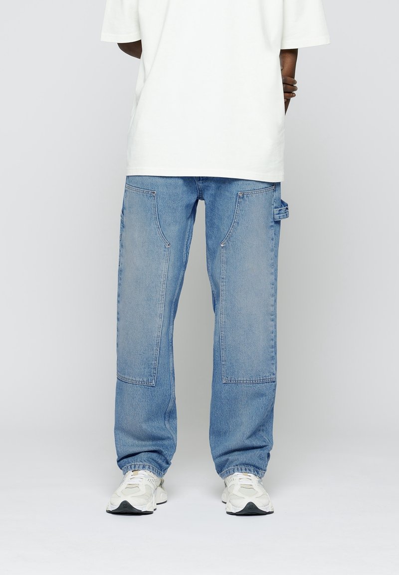 Jean large en denim bleu clair avec de grandes poches avant, coutures renforcées et une étiquette sur le côté. Porté avec un t-shirt blanc oversized.