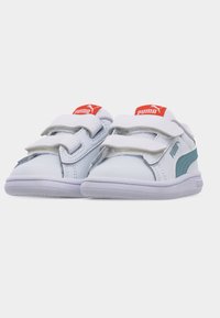 Λευκά παιδικά αθλητικά παπούτσια Puma με τυρκουάζ ρίγα, κόκκινες ετικέτες με το λογότυπο και δύο λευκές λουρίδες Velcro σε κάθε παπούτσι, παρουσιάζονται σε λευκό φόντο.