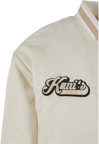 Beige Bomberjacke mit einem gerippten Kragen. Auf der linken Seite mit schwarzem und goldenem gesticktem Text "Kani's Diner". Glatte Textur.
