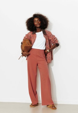 Femme aux cheveux bouclés portant un pantalon large couleur rouille, chemise blanche, veste fleurie, tenant un sac en daim marron et des sandales, debout contre un mur blanc.
