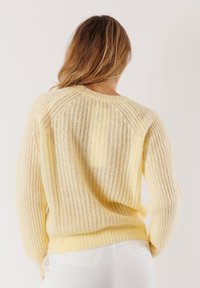Donna con lunghi capelli che indossa un maglione lavorato a maglia giallo chiaro e pantaloni bianchi, vista da dietro su uno sfondo semplice.