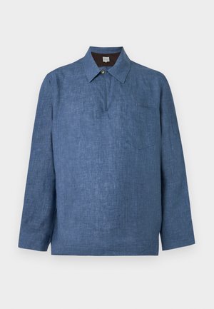 Homme debout, main dans la poche, portant une chemise bleue ample à col, un jean clair à jambes larges, une ceinture marron et des mocassins noirs sur fond blanc.