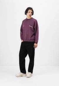 Fialová mikina s okrúhlym výstrihom a voľným strihom. Čierne široké nohavice a béžové tenisky dopĺňajú outfit. Minimalistický dizajn s detailom loga.