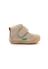 Botín de ante beige con una correa de Velcro, forma redondeada y suela de goma marrón claro. Presenta costuras en contraste y un pequeño logo verde.