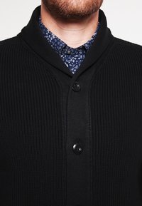 Schwarze Strickjacke mit strukturierter Oberfläche, ausgestattet mit einem Schalkragen und drei schwarzen Knöpfen, über einem blauen Hemd mit floralem Muster getragen.