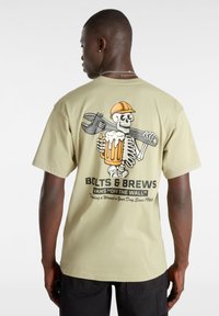 Camiseta de algodón verde oliva claro con un gráfico de un esqueleto sosteniendo una llave inglesa y una cerveza, con el texto "Bolts & Brews" debajo del diseño.