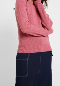 Maglione rosa lavorato a maglia con orlo e polsini a coste, caratterizzato da una vestibilità comoda. Indossato con pantaloni blu scuro con cuciture bianche a contrasto.