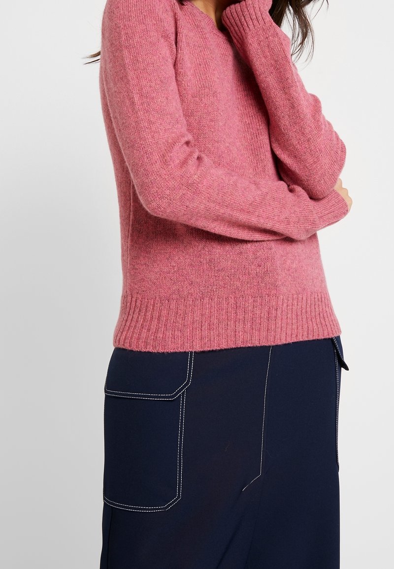 Maglione rosa lavorato a maglia con orlo e polsini a coste, caratterizzato da una vestibilità comoda. Indossato con pantaloni blu scuro con cuciture bianche a contrasto.