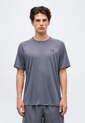 Jonge man staat stil, draagt een effen grijs Under Armour sportshirt met korte mouwen en bijpassende korte broek tegen een effen achtergrond.
