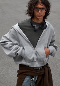 Grå zip-up hoodie med oversized ærmer, parret med en mørkegrøn t-shirt. Sorte hovedtelefoner hviler om halsen, iført blå jeans.