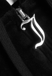 Lesklý stříbrný jezdec zipu ve tvaru stylizovaného plamene připevněný k černému texturovanému zipu.