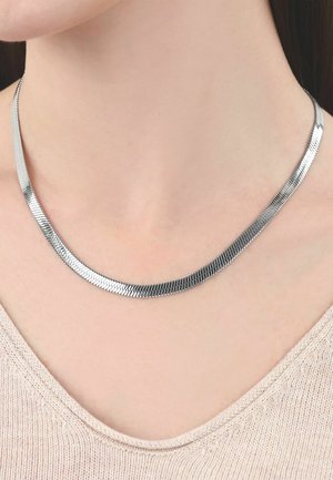 Zilveren platte vissengraatketting gedragen op een blote hals boven een beige gebreide top met een brede halslijn.