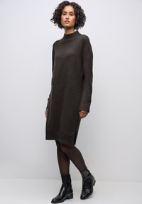 Strickbraunes Kleid mit hohem Kragen, langen Ärmeln, seitlichen Schlitzen und lässiger Passform, kombiniert mit schwarzen Knöchelstiefeln.