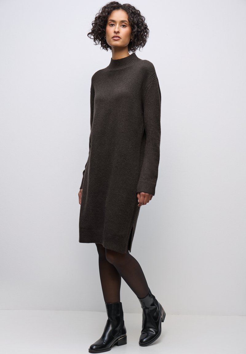 Strickbraunes Kleid mit hohem Kragen, langen Ärmeln, seitlichen Schlitzen und lässiger Passform, kombiniert mit schwarzen Knöchelstiefeln.