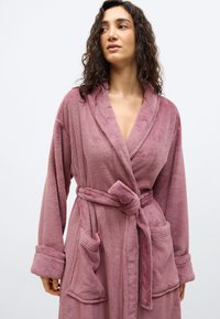 Γυναίκα που φοράει ένα απαλό ρόμπα σε χρώμα μωβ, με δεμένη ζώνη και δύο μπροστινές τσέπες, κοιτάζοντας ελαφρώς προς τα πλάγια σε απλό φόντο.