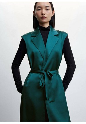 Femme aux cheveux raides et foncés portant un gilet en satin vert foncé sans manches par-dessus un haut noir à manches longues, debout les mains dans les poches.