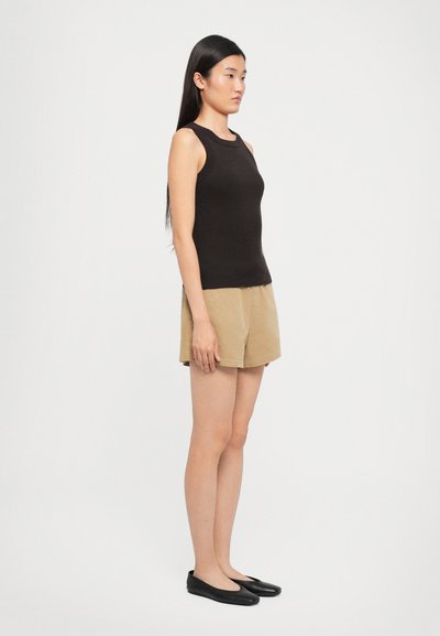 Black ribbed tank top, tan shorts, and black flats. Model stands sideways, showing fitted design and sleeveless style. Smooth textures.