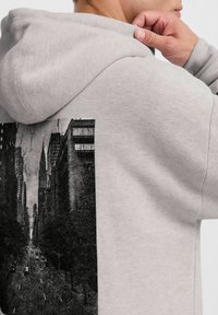 Sweat-shirt à capuche gris avec une impression en noir et blanc de paysage urbain au dos. Tissu doux avec une coupe décontractée et des poignets côtelés.
